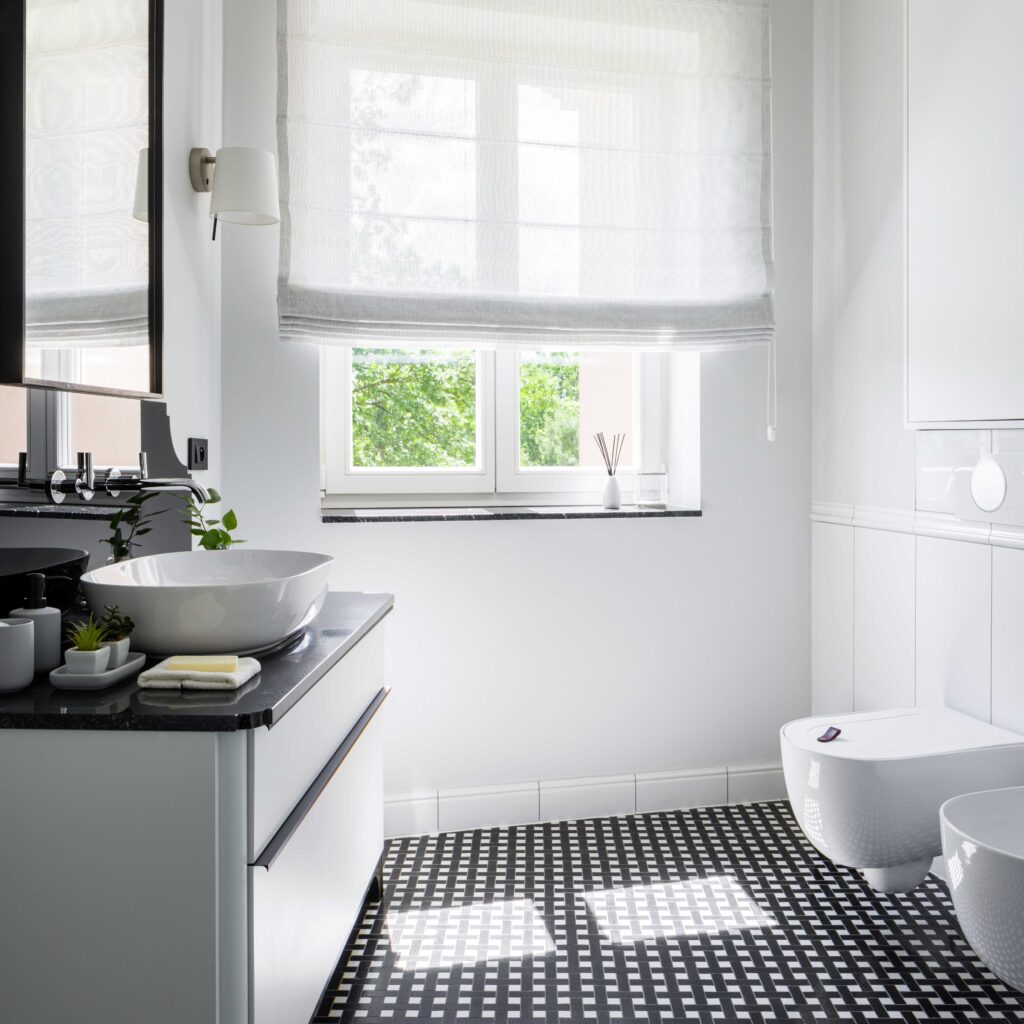 Hell gestaltetes Badezimmer mit einem großen Fenster, das viel natürliches Licht hereinlässt, ausgestattet mit einem modernen Waschbecken und einer Toilette, schwarz-weiße Bodenfliesen vervollständigen das schicke Design