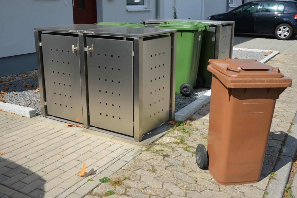 Edelstahl Mülltonnenbox für vier Mülltonnen, die zwei grüne und eine braune Mülltonne sauber und ordentlich auf einem gepflasterten Bereich aufbewahrt.