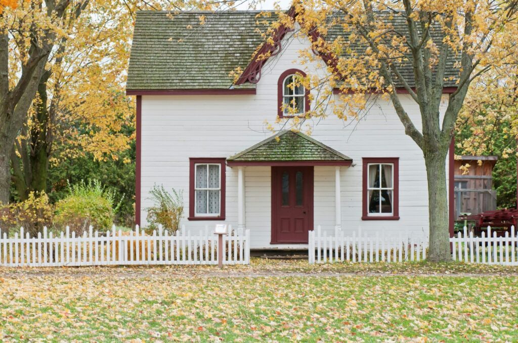 Weiß gestrichenes Haus mit kleinem Gartenzaun im Herbst, umgeben von buntem Laub. Weißes Landhaus mit roter Tür und kleinem weißen Zaun, Herbstlaub im Vordergrund.